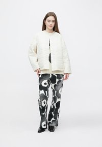 Gesteppte cremefarbene Jacke mit rundem Ausschnitt, kombiniert mit schwarz-weiß gemusterten Hosen mit Blumenmuster und schwarzen Schuhen.