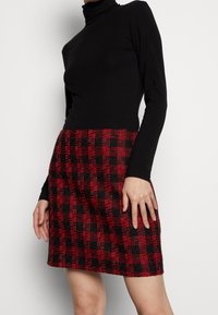 Maglione a collo alto nero abbinato a una mini gonna a quadri rossa e nera, realizzata in tessuto testurizzato con una forma strutturata.