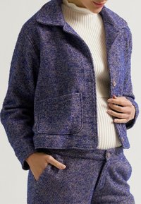 Gewebte Hahnentrittmuster-Jacke in Blau und Weiß mit einem Kragenhalsausschnitt, einer Fronttasche und einer Knopfleiste. Die strukturierte Oberfläche verleiht Tiefe.