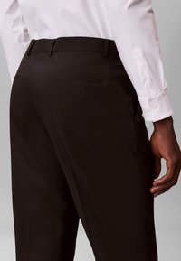 Άνδρας που φοράει μαύρο παντελόνι φόρμας και λευκό πουκάμισο με μακριά μανίκια και κουμπιά, με το δεξί χέρι χαλαρό στο πλάι.