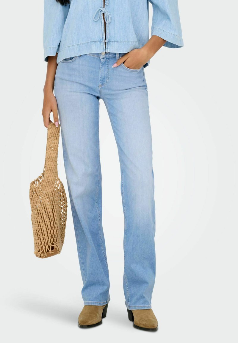 Mujer con jeans de mezclilla azul claro y chaqueta a juego, sosteniendo un bolso de mimbre beige y usando botas de tacón de ante color beige.