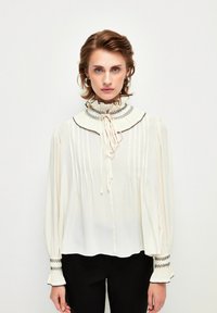 Weiße Bluse mit hohem, rüschendem Kragen, gefalteter Front und schwarzem Besatz. Lange Ärmel mit dekorativem gesticktem Bündchen. Weicher, leichter Stoff.