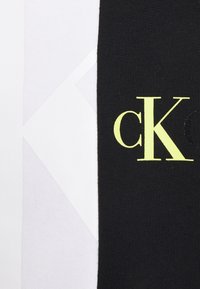 Tessuto nero con finitura testurizzata, caratterizzato dal luminoso logo "cK" giallo e sezioni adiacenti di bianco e grigio chiaro in tessuto liscio.