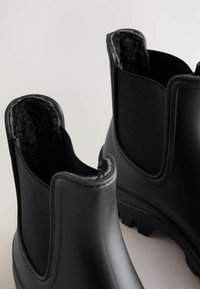Schwarze Gummistiefel bis zum Knöchel mit strukturiertem Innenfutter und elastischen Seitenpaneelen. Profilierte Sohlen für besseren Halt. Glatte Oberfläche ohne sichtbare Markierungen oder Logos.