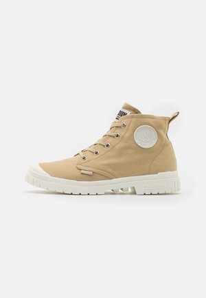 Beige canvas ankle boots with white rubber soles, lace-up design, and a round toe. Features a circular logo patch on the side.