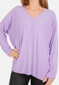 Helllila Langarmoberteil mit V-Ausschnitt, lockerer Passform und weicher Textur. Endet an den Hüften, getragen über schwarzen Hosen.