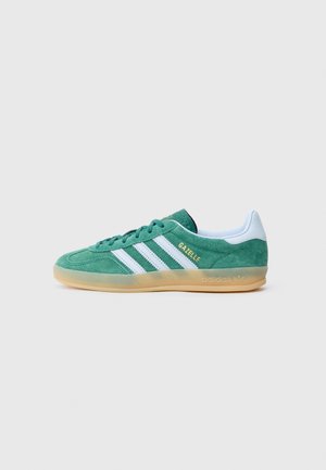 Grøn Adidas Gazelle sneaker med hvide striber, hvid hælflap, gummisål og gylden "GAZELLE" tekst på siden.