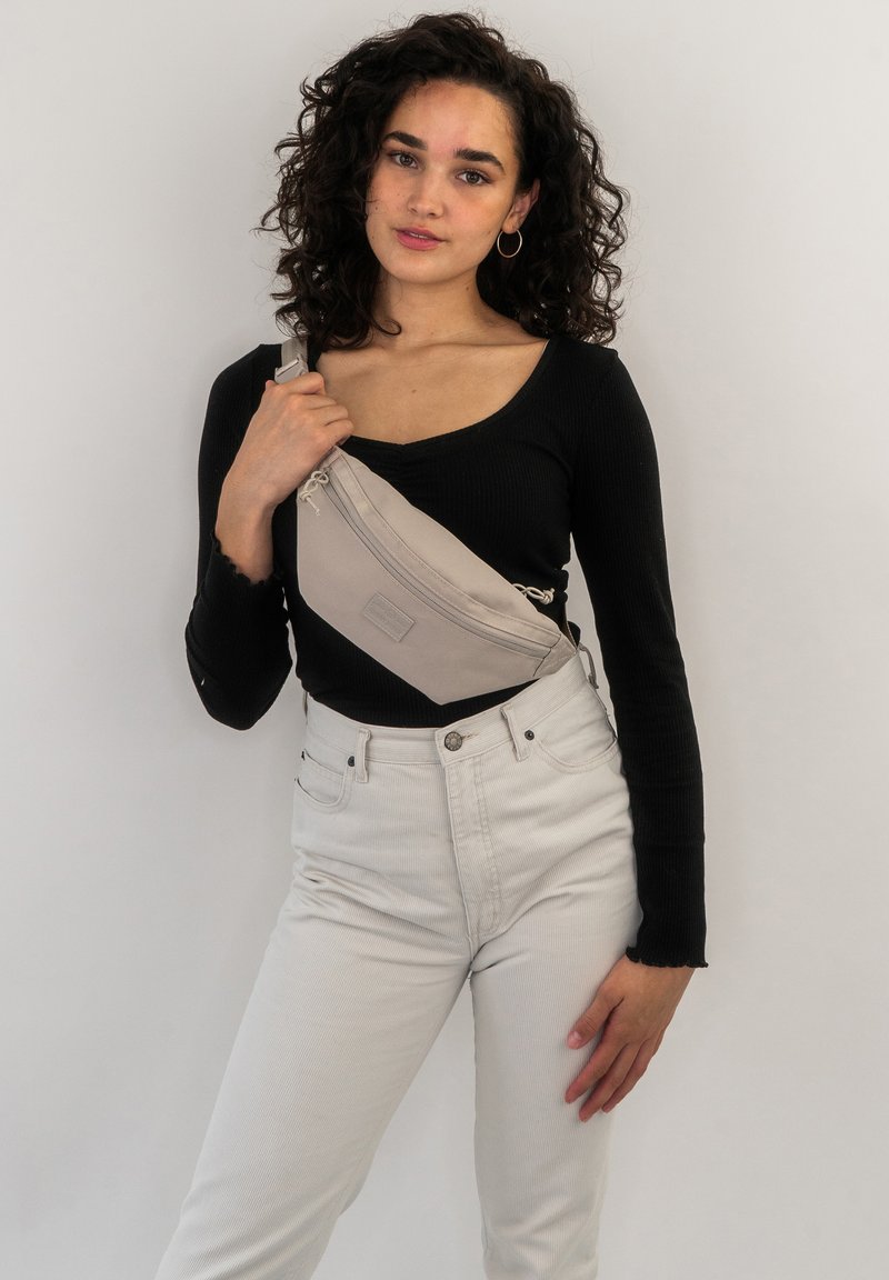 Sac bandoulière beige en tissu, avec une poche zippée inclinée et une sangle ajustable. Le modèle porte un haut noir et un jean clair.