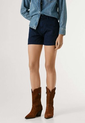 Persona che indossa pantaloncini blu scuro a vita alta, una camicia di jeans e stivali da cowboy in suede marrone fino a metà polpaccio, in piedi davanti a uno sfondo semplice.