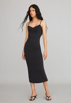 Donna con lunghi capelli scuri che indossa un vestito midi nero aderente con sottili spalline e sandali neri con tacco, in piedi davanti a uno sfondo grigio semplice.