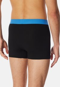 Uomo che indossa boxer neri con una cintura blu, in piedi con le braccia rilassate ai lati, visto da dietro su uno sfondo bianco.