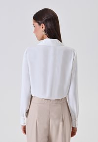 Camicia bianca a maniche lunghe con colletto, caratterizzata da una texture liscia e polsini con bottoni. Abbinata a pantaloni beige a vita alta.