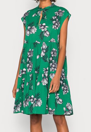 Zielona sukienka bez rękawów z białym i czarnym kwiatowym nadrukiem, piętrową spódnicą oraz małym wycięciem z przodu przy dekolcie, noszona przez stojącą osobę.