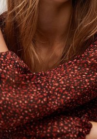 Gros plan d'une personne aux cheveux châtain clair portant un chemisier à manches longues à motifs rouge foncé et noir, bras croisés près de la poitrine.