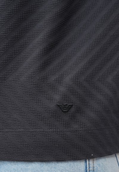 Čierna textúrovaná látka s vzorom rybej kosti obsahuje malé tmavé logo. Oblečenie je doplnené svetlým džínsovým materiálom v dolnej časti.