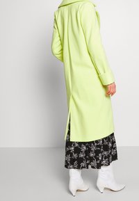 Bright lime green long coat with a wide collar, side slits, and rolled cuffs, worn over a black floral patterned skirt and white ankle boots.