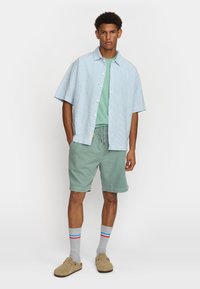 Camicetta a righe azzurro chiaro a maniche corte, T-shirt verde, pantaloni corti menta, calze grigie con strisce rosse e blu, e scarpe beige senza lacci.