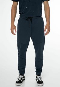 Marineblaue Jogginghose im tapered Fit, mit einem Kordelzugbund, seitlichen Einsätzen und weichem Material. Kombiniert mit schwarzen Turnschuhen.