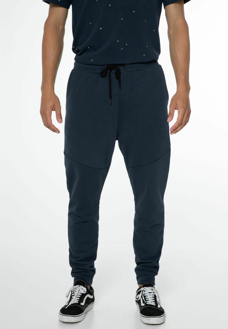 Marineblaue Jogginghose im tapered Fit, mit einem Kordelzugbund, seitlichen Einsätzen und weichem Material. Kombiniert mit schwarzen Turnschuhen.