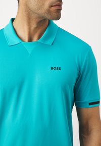 Turkizna polo majica iz teksturiranega blaga, z črnim logotipom "BOSS" na prsih in črnimi detajli na rokavih.