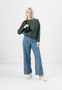 Dark green sweatshirt, cropped design, wide blue denim jeans, white sneakers. Model holds a small black handbag. Minimalistic background.