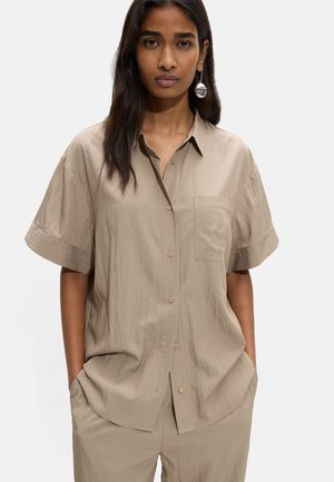 Kobieta ubrana w luźną beżową koszulę na krótki rękaw z guzikami oraz pasujące spodnie, z rękami w kieszeniach na jednolitym tle.