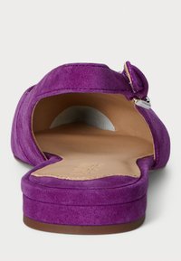 Fioletowe zamszowe buty z odkrytymi plecami, z okrągłym czubkiem, płaską podeszwą i ozdobnym detalem w postaci kokardy na pasku. Miękka tekstura i gładkie wnętrze.