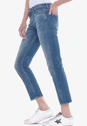 Denim jeans in mid-blue, slim-fit design with slight fading, five-pocket styling, and button closure. Styled with white sneakers.