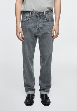 Mann trägt graue Jeans mit geradem Bein und umgeschlagenen Säumen, schwarze Lederschuhe und ein hellgraues T-Shirt, das in die Hose gesteckt ist, und steht vor einem einfachen Hintergrund.