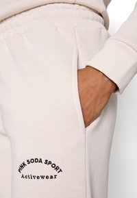 Ljusbeige träningsoverallbyxor med avslappnad passform, som har en sidoficka, ribbade mudd och en svart broderad logotyp.