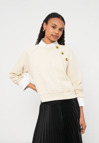 Μπεζ φούτερ με στρογγυλή λαιμόκοψη, διακοσμημένο με τρία χρυσά κουμπιά στον αριστερό ώμο και μακριά μανίκια πάνω από ένα λευκό πουκάμισο με γιακά.