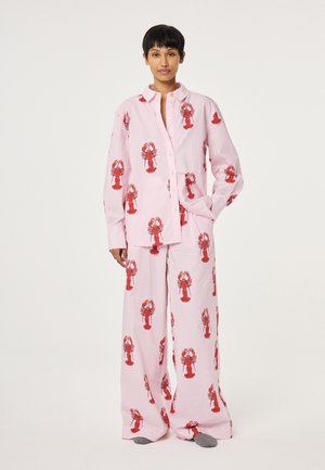 Person trägt lockeres rosa Pyjama-Set mit roten Hummerdruck, steht mit einer Hand in der Tasche vor einem einfarbigen Hintergrund.