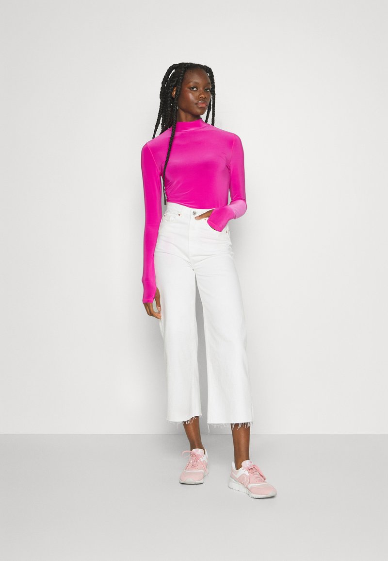 Femme avec de longues tresses portant un haut à manches longues rose vif, un pantalon large blanc court et des baskets roses, se tenant devant un fond uni.