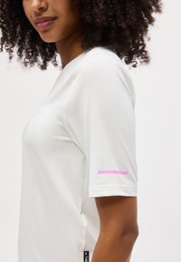 Γυναίκα με σγουρά μαλλιά που φοράει λευκό πουκάμισο με κοντά μανίκια, εμφανίζοντας μια ροζ ετικέτα "50+ Προστασία από την Υπεριώδη Ακτινοβολία" στο μανίκι.