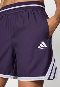 Pantaloni sportivi viola con vita in rete, tre strisce bianche di lato e un testo bianco con la scritta "BASKETBALL."