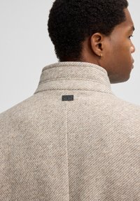 Lichtbeige wollen blend jas met een hoge kraag en diagonale visbroodpatroon. Bevat een klein zwart label bij de hals.