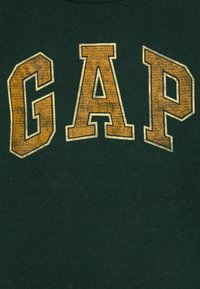 T-shirt verde escuro com "GAP" impresso em letras grandes e desgastadas de cor dourada. Decote redondo, textura suave, design casual.