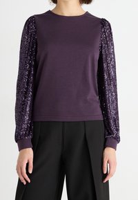 Top de manga larga morado con cuerpo de tela suave y mangas globo adornadas con lentejuelas. Los puños acanalados completan el diseño. Combinado con pantalones negros.