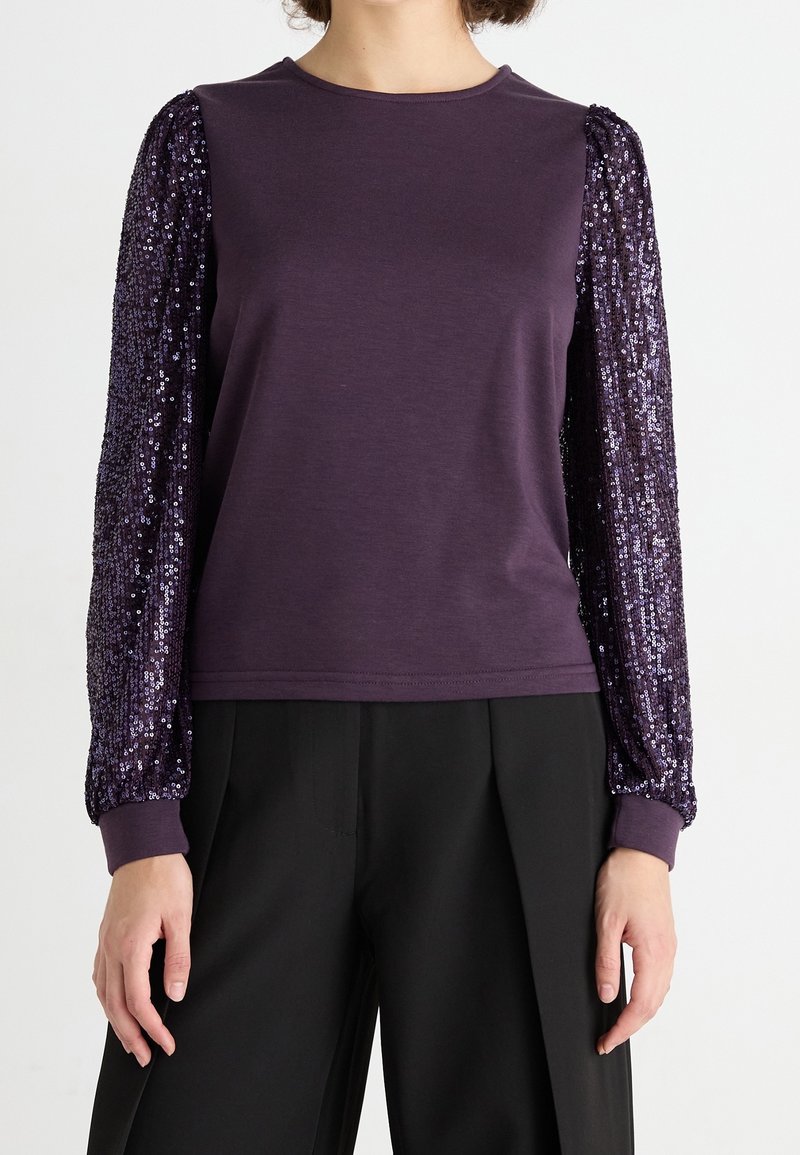 Top de manga larga morado con cuerpo de tela suave y mangas globo adornadas con lentejuelas. Los puños acanalados completan el diseño. Combinado con pantalones negros.