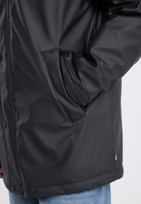Black waterproof jacket featuring a smooth texture, snap buttons, and side pockets. Slight sheen, with a logo tag at the hem.