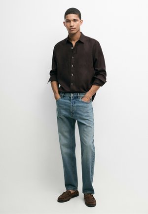 Giovane uomo in piedi con le mani nelle tasche, indossa una camicia marrone scuro bottonata, jeans azzurri e mocassini marroni su sfondo bianco.