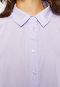 Světle fialová košile s límečkem a zapínáním na knoflíky, s hladkým povrchem a klasickým designem. Knoflíky jsou z průhledného plastu.