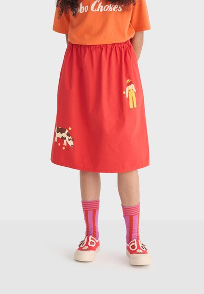 Enfant portant une jupe rouge avec des imprimés d'agriculteur et de vache, un t-shirt orange, des chaussettes rayées roses et des chaussures rouges avec des détails floraux, debout devant un fond uni.