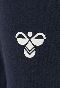 Marineblåt stof prydes af et hvidt logo designet som en stiliseret bi, med buede vinger og et chevronmønster nedenunder, på en tekstureret overflade.