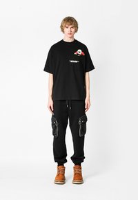 Camiseta negra de gran tamaño con bordado floral y texto, combinada con pantalones cargo negros con bolsillos con cremallera y herrajes plateados. Footwear marrón.