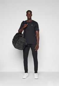 Zwarte sport T-shirt met logo, bijpassende zwarte sweatpants, witte sneakers en een zwarte sporttas met gaasaccenten.