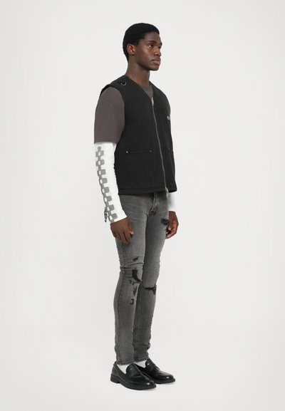 Vest negru fără mâneci cu fermoar, asortat cu un tricou gri și o bluză albă cu mânecă lungă, având un accent cu model. Blugi gri decolorati și ciobiti, și pantofi negri.