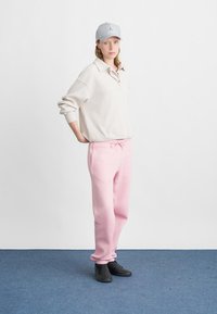 Pulover ribbed de culoare bej deschis cu guler, asociat cu pantaloni de trening roz, dotati cu un cordon ajustabil. Pantofi negri, pălărie gri.