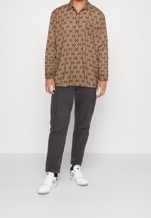 Man draagt een bruin patroonachtig shirt met lange mouwen, donkergrijze cargobroek en witte sneakers, staand tegen een egale achtergrond.