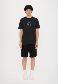 Sort kortærmet T-shirt med et hvidt rundt logo på brystet, parret med sorte shorts og hvide sneakers. Minimalistisk design, almindeligt bomuldsstof.
